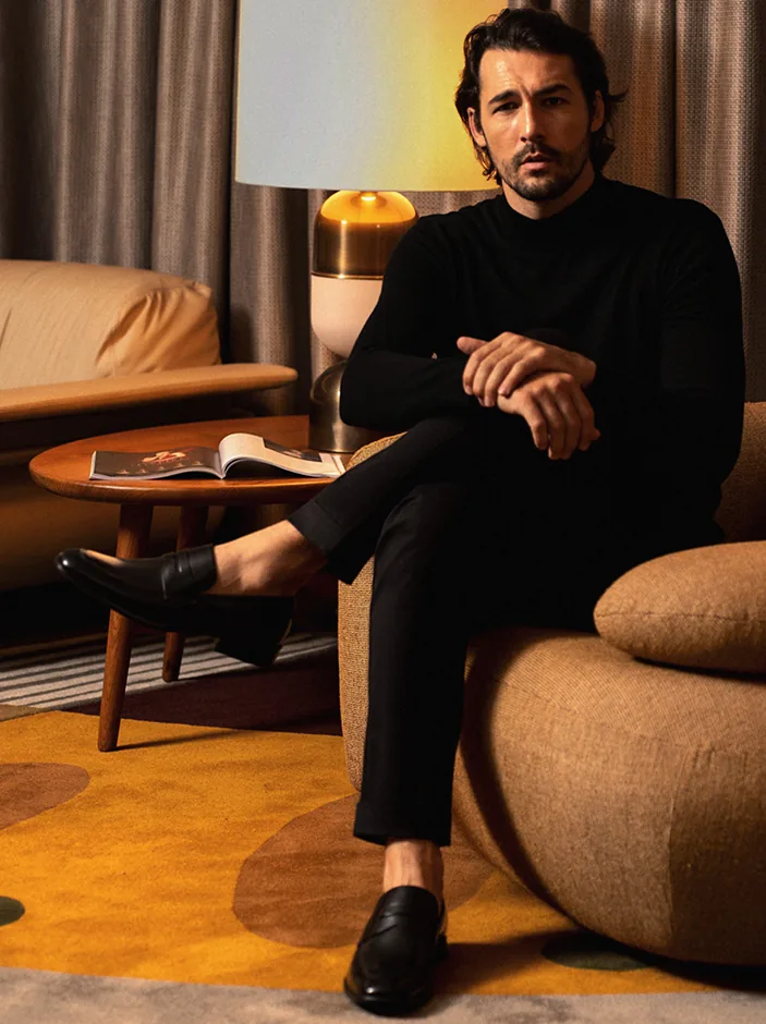 Homem de look completo, já calçado, sentado em ambiente de luz quente.